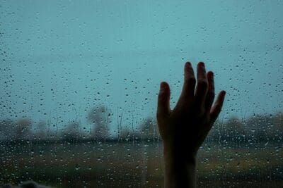 A hand pressed to a rain-streaked window in blue-grey light, echoing a widow's quiet grief