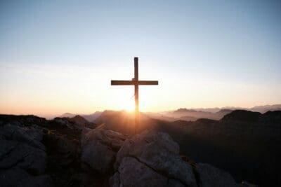 A wooden cross silhouetted on a mountaintop at sunrise, light breaking behind the beam