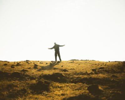 A figure stands arms open in a golden field, small before the wide sky, a picture of dependence