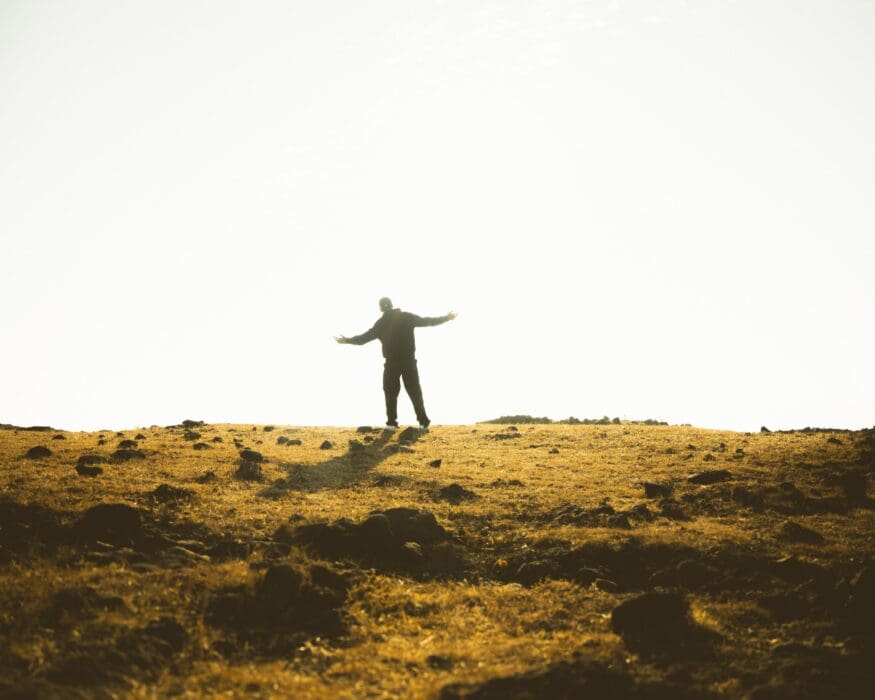 A figure stands arms open in a golden field, small before the wide sky, a picture of dependence