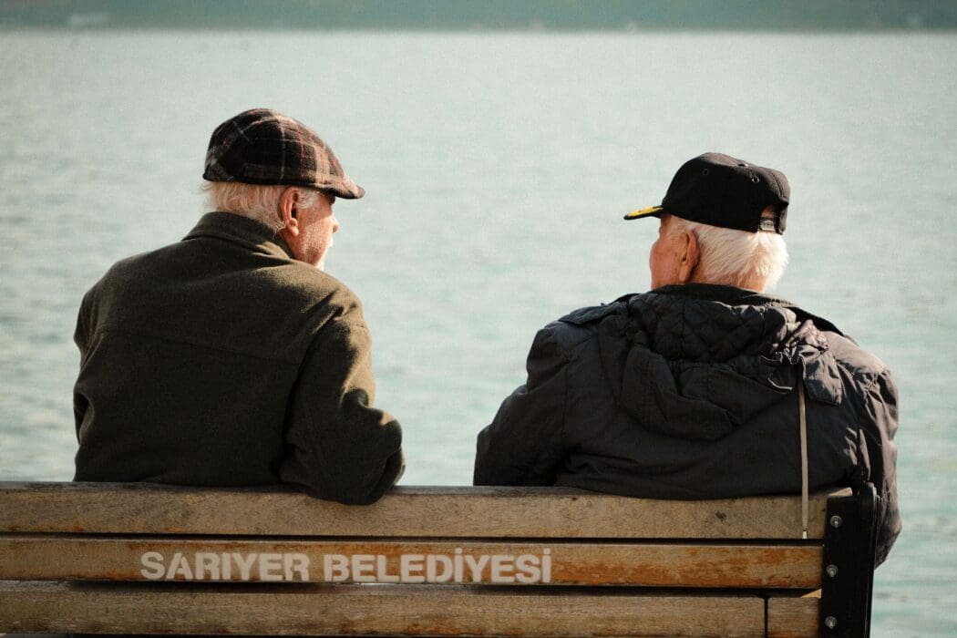 Two old friends sit side by side on a bench facing the water, a picture of good news shared or kept