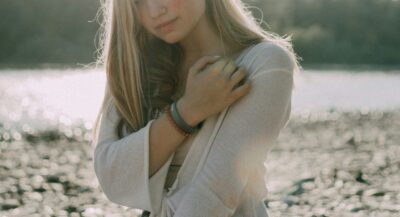 A young woman by the water in soft light, hand to her shoulder, weighing what her heart is telling her.