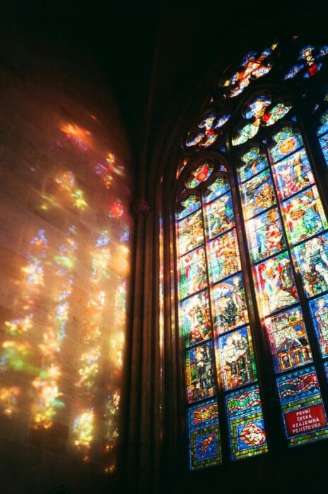 Stained glass casting colorful light across a stone cathedral wall, the setting of the Pastor John parable.