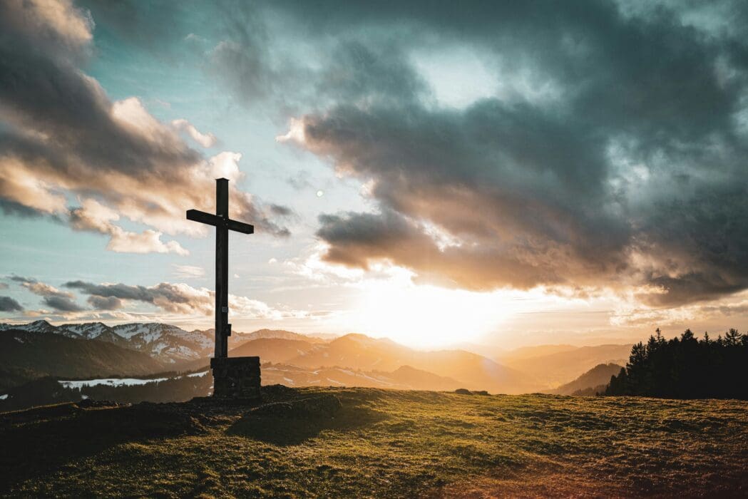 A wooden cross on a mountain summit under a dramatic sunset sky, sunbeams breaking through the clouds.