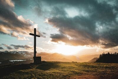 A wooden cross on a mountain summit under a dramatic sunset sky, sunbeams breaking through the clouds.