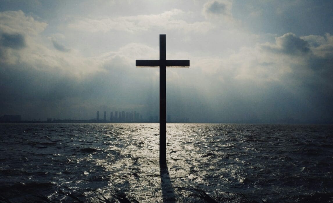 A dark cross standing in choppy ocean water with light breaking through storm clouds above a skyline.