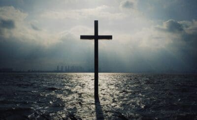 A dark cross standing in choppy ocean water with light breaking through storm clouds above a skyline.
