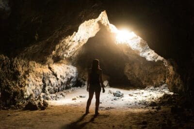 A woman silhouetted in a dark cave as bright light streams through an opening above her.